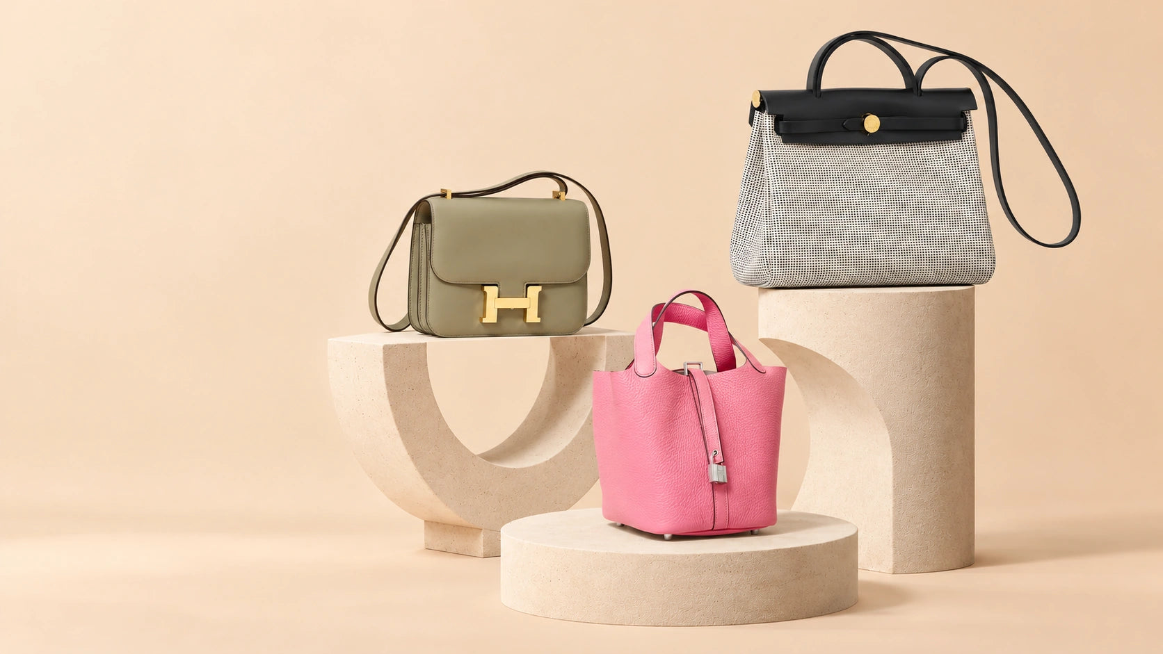 Olive Hermès Mini Constance with gold hardware, black-and-white Herbag 31 and rose-pink Picotin 18 on cream sculptural stone plinths against a warm beige studio backdrop
